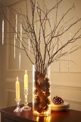 Eine Vase ist gefüllt mit Tannenzapfen und einer Lichterkette. In der Vase stehen außerdem auch noch ein paar Zweige, die mit silberner Weihnachtsdeko behangen sind.