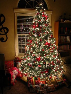 Ein hell erleuchteter Weihnachtsbaum, unter dem schon Geschenke liegen steht in einem schummrigen Wohnzimmer.