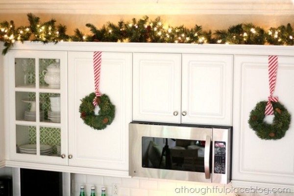 Auf einem weißen Küchenschrank liegen grüne Tannenzweige, die mit einer Lichterkette erleuchtet werden. 