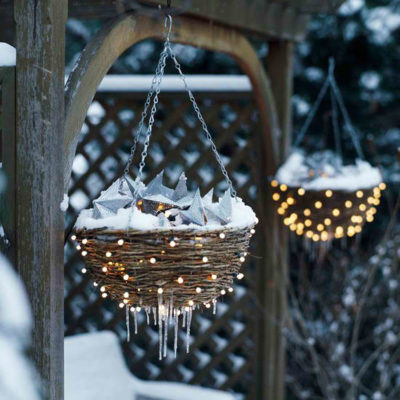 zwei hängende Körbchen im Garten, schneebedeckt und durch kleine LEDs sanft leuchtend