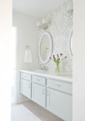 Ein weißer Waschtisch vor einer weiß gefliesten Wand, darüber ein weißer, ovaler Spiegel mit Wandlampen, die weiße Stoffschirme haben. Der einzige Farbklecks ist ein Strauß pinker Tulpen auf dem Waschtisch.