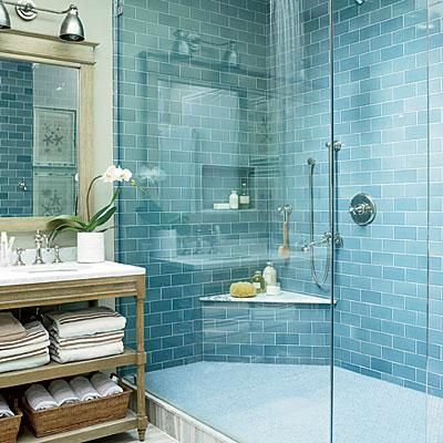 Man sieht eine blau geflieste Wand in einer Duschkabine aus Glas, daneben steht ein hölzerner Waschtisch mit Spiegel, über dem metallische Industrielampen hängen.