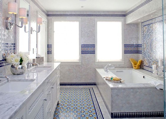 in einem opulenten Badezimmer mit blauen Mosaikfliesen steht links der Waschtisch, rechts die Badewanne. Als Spiegelleuchten beim Waschtbecken dienen zwei Wandleuchten mit rosafarbenen Schirmen.