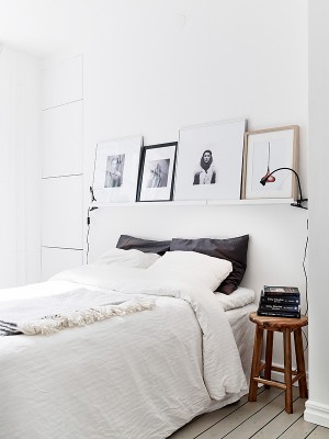 Ein weißes Schlafzimmer mit einem großen Doppelbett, darüber eine Bilderleiste, an der zwei schwarze Klemmleuchten mit Schwanenhals hängen, ist zu sehen.