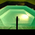 Ein sechseckiger Pool mit einer umlaufenden Stufe ist mit einer Unterwasserbeleuchtung ausgestattet, die das Wasser im Dunklen türkis wirken lässt.