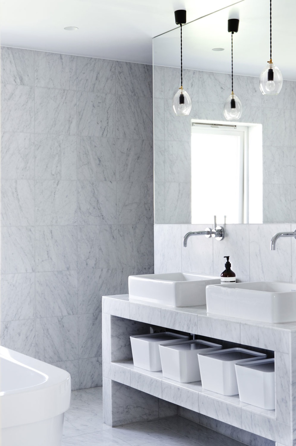Ein hell gefliestes Bad mit einem eleganten Waschtisch aus Stein, vor den Spiegeln hängen zwei Deckenleuchten.