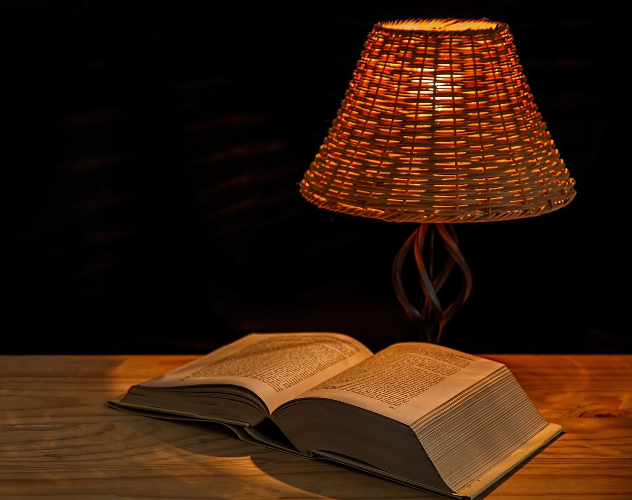 Auf einem Tisch liegt ein dickes, aufgeschlagenes Buch, daneben steht eine Tischleuchte mit einem Lampenschirm aus Korbgeflecht. Das Licht ist sehr warm.
