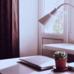 Eine weiße Schreibtischlampe steht vor einem Fenster auf einem Tisch, daneben ein Laptop und eine Pflanze.