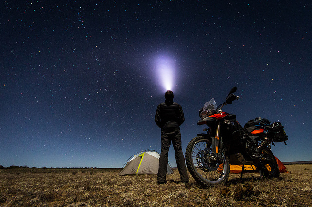 Eine Person steht nachts mit Zelt und Mototrrad in der freien Natur und schaut nach oben, eine Stirnlampe leuchtet in den Himmel
