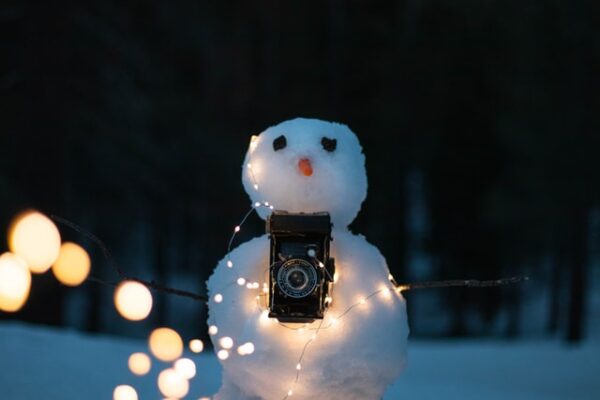 Ein Schneemann hält eine Kamera in der Hand un wurde mit einer Lichterkette umwickelt