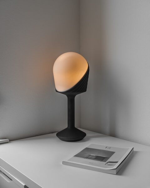 Auf einem Schreibtisch in einer Ecke steht eine schwarze, moderne Tischlampe mit kunstvollem Milchglasschirm