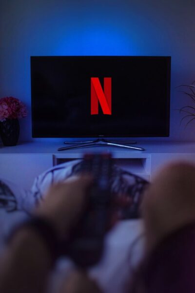 Jemand hält eine Fernbedienung vor einen Fernseher, auf dem das Netflixlogo zu sehen ist. Dahinter ist eine blaue Hintergrundbeleuchtung.