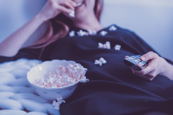 Eine Frau liegt lächelnd auf der Couch und isst Popcorn. Sie hat eine Fernbedienung in der Hand.